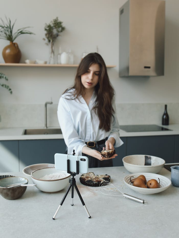 Woman creating a cooking blog video in a modern kitchen with phone on tripod.