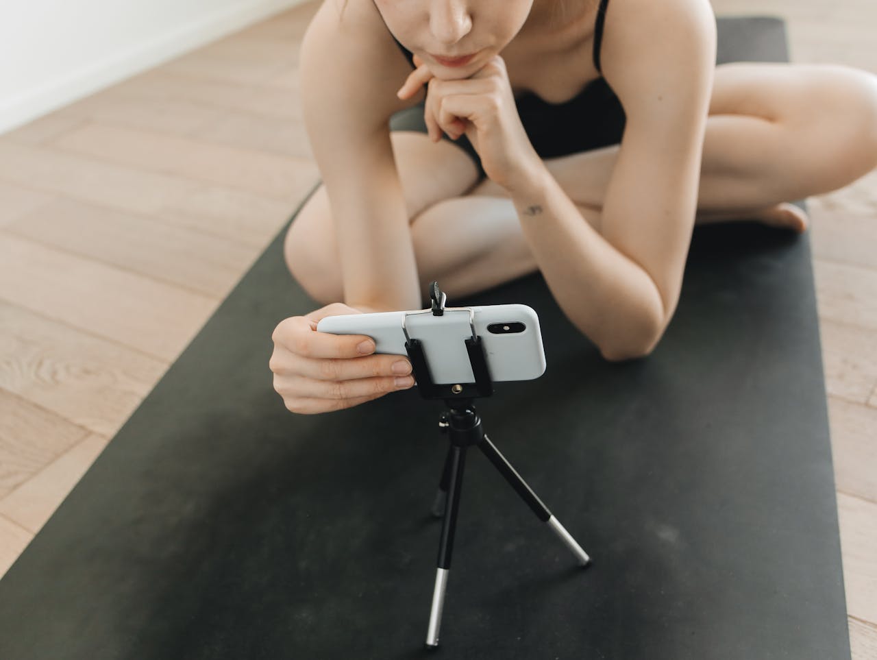 A woman in activewear vlogs using a smartphone and tripod while seated on a yoga mat indoors.